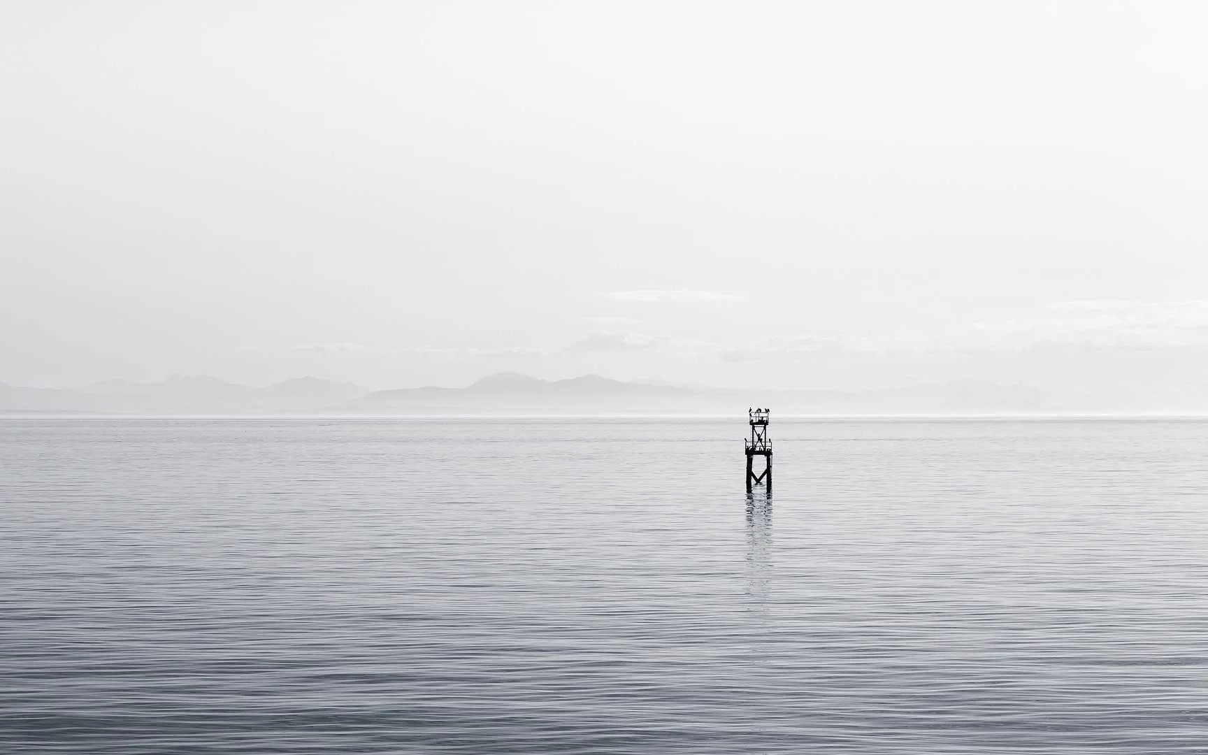 A lone marker post standing in flat, silver water under a white sky.
