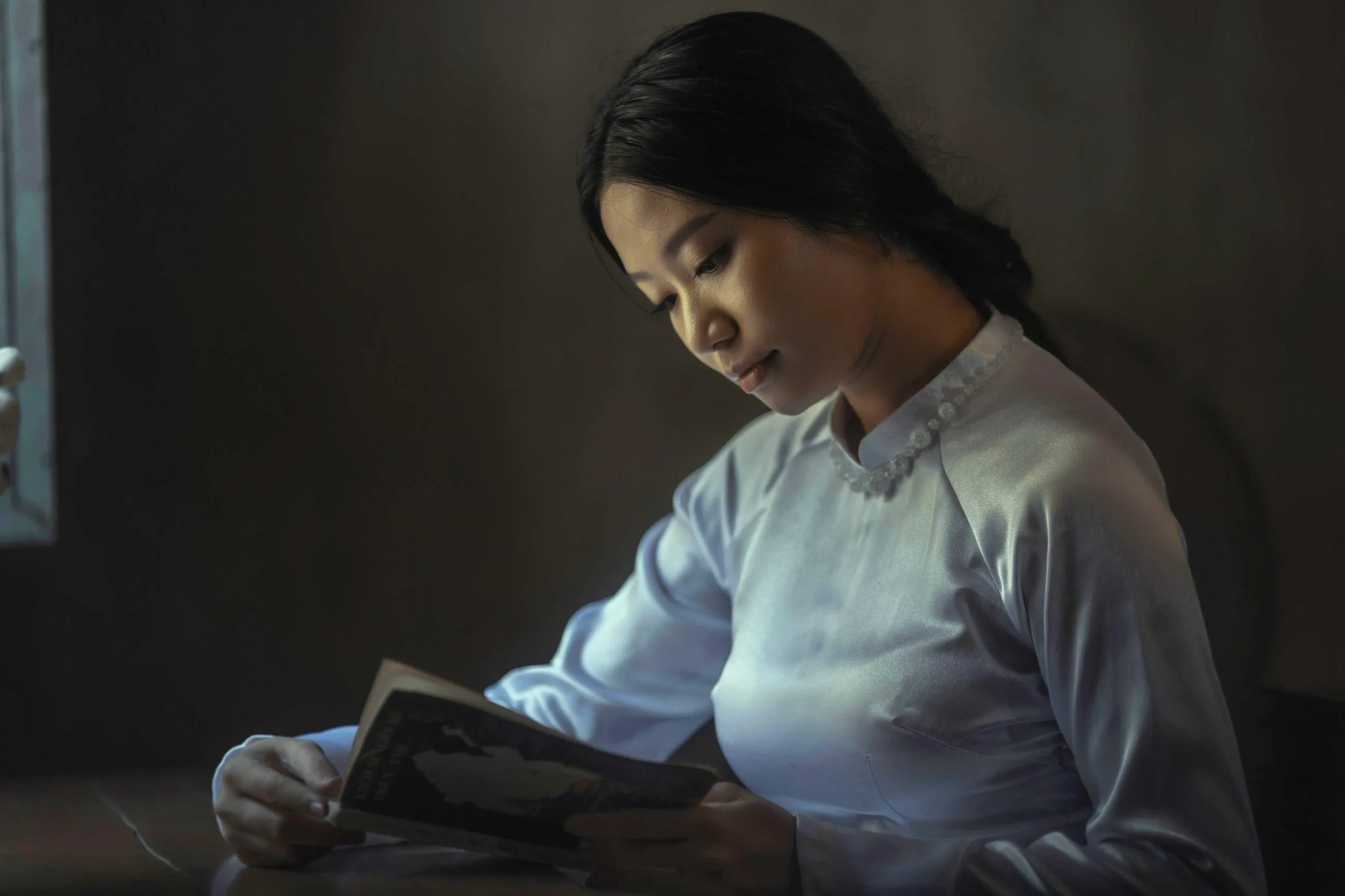 A woman in a pale blue tunic reads by window light in a dark room.