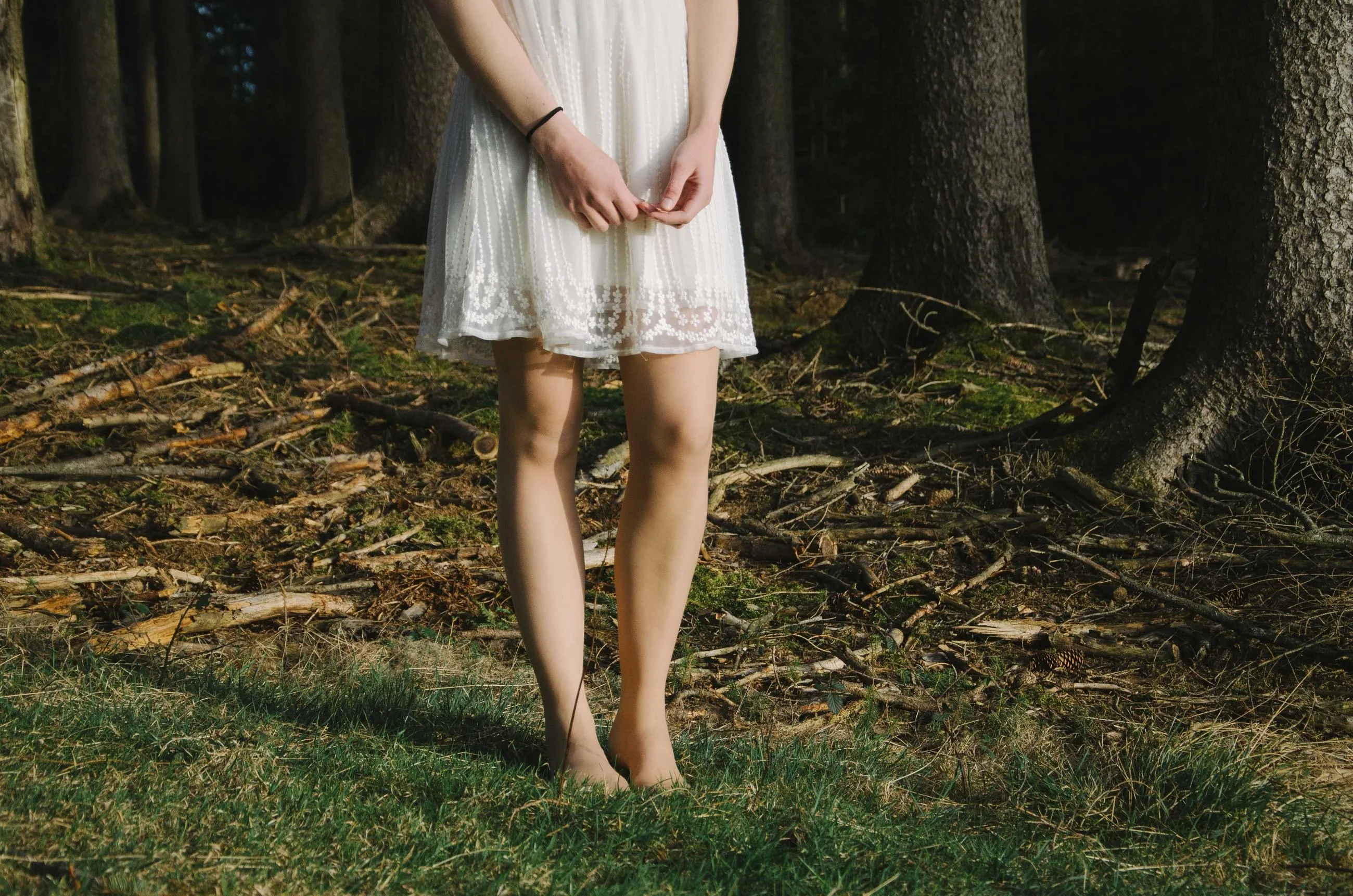 Bare legs and a white lace dress against the dark floor of a pine forest.