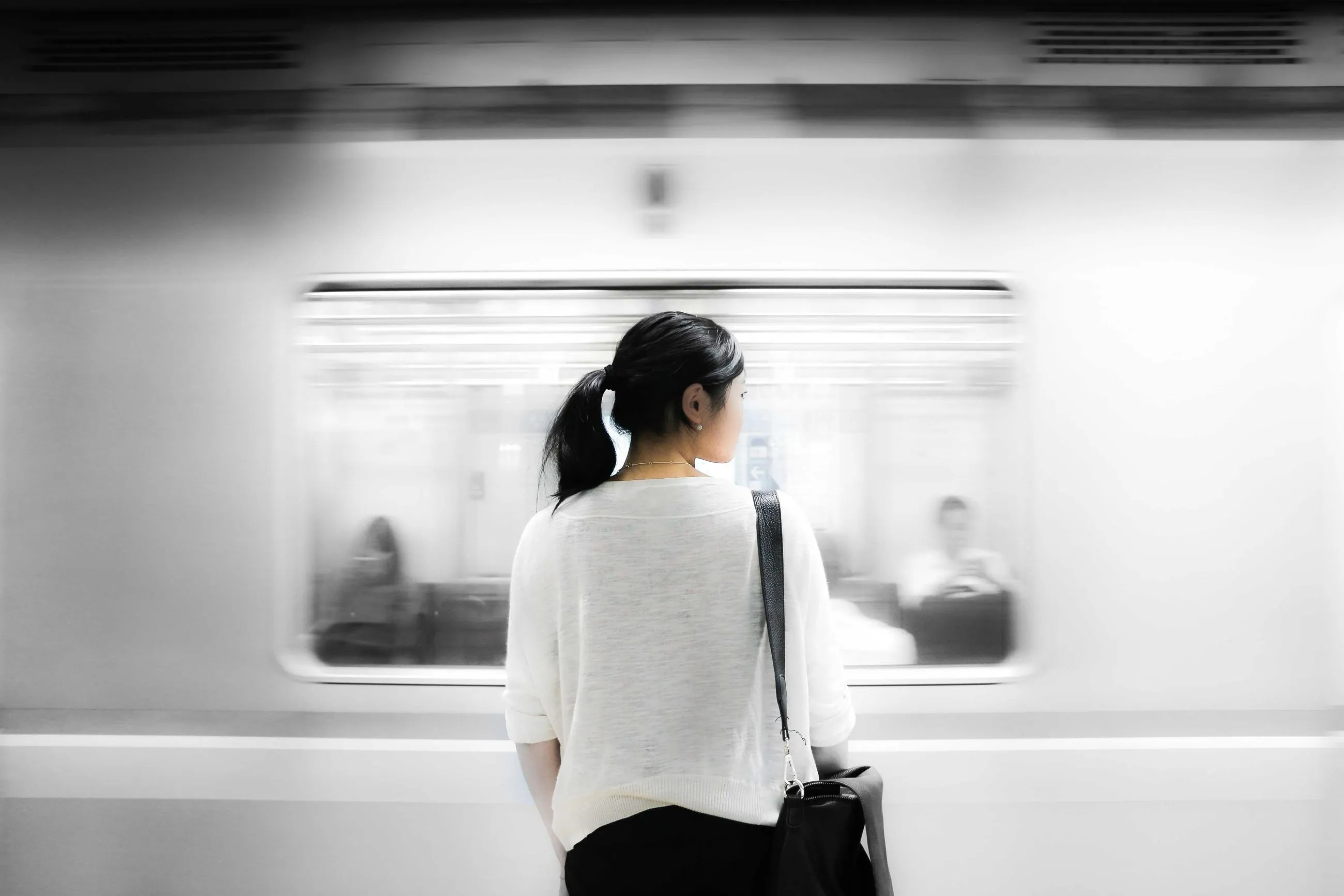 A woman with a ponytail waits on a white subway platform as a train streaks past.