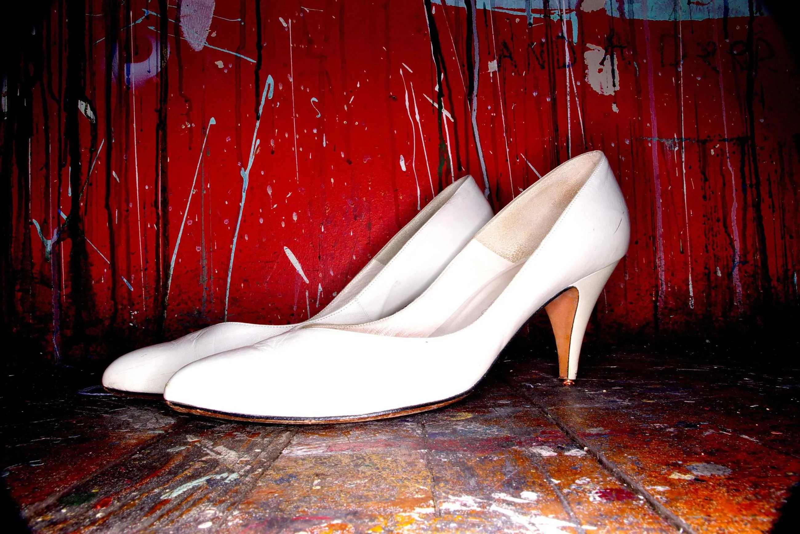 A pair of white leather court shoes on a paint-spattered stage in front of a red wall.
