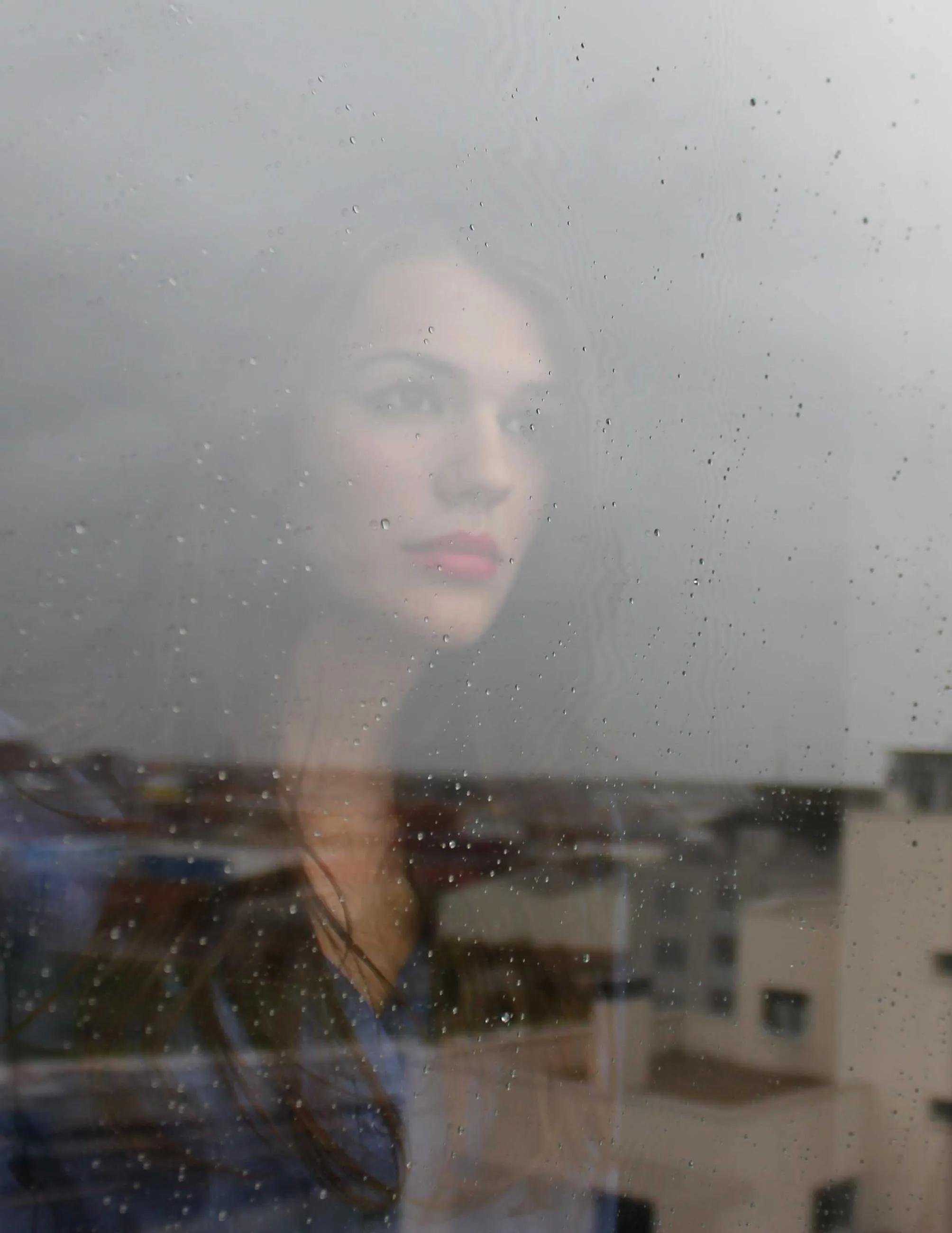 A woman’s face reflected in a rain-marked window over a city of low buildings.
