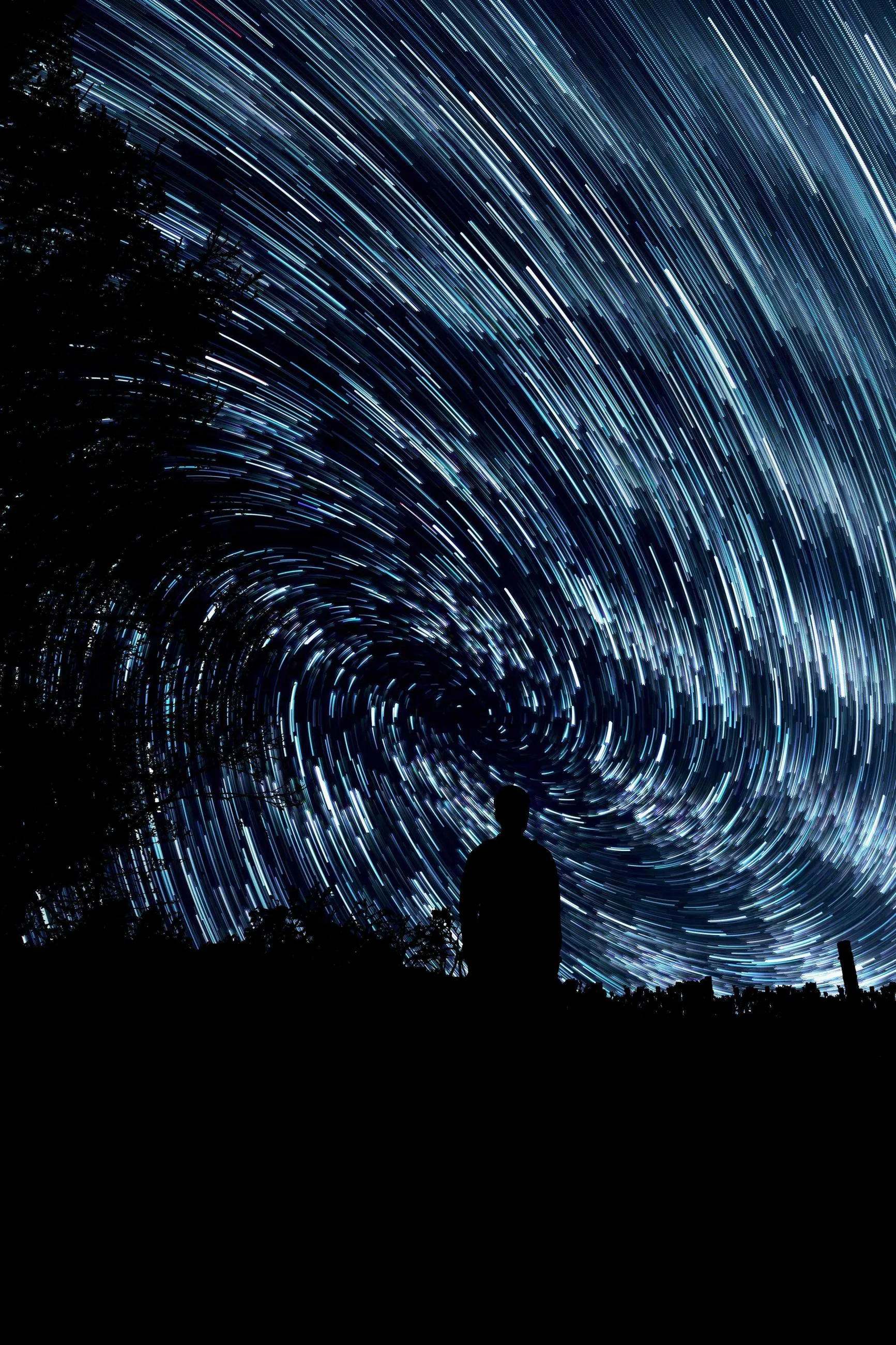 Star trails arc above the silhouette of a person and a single tree.