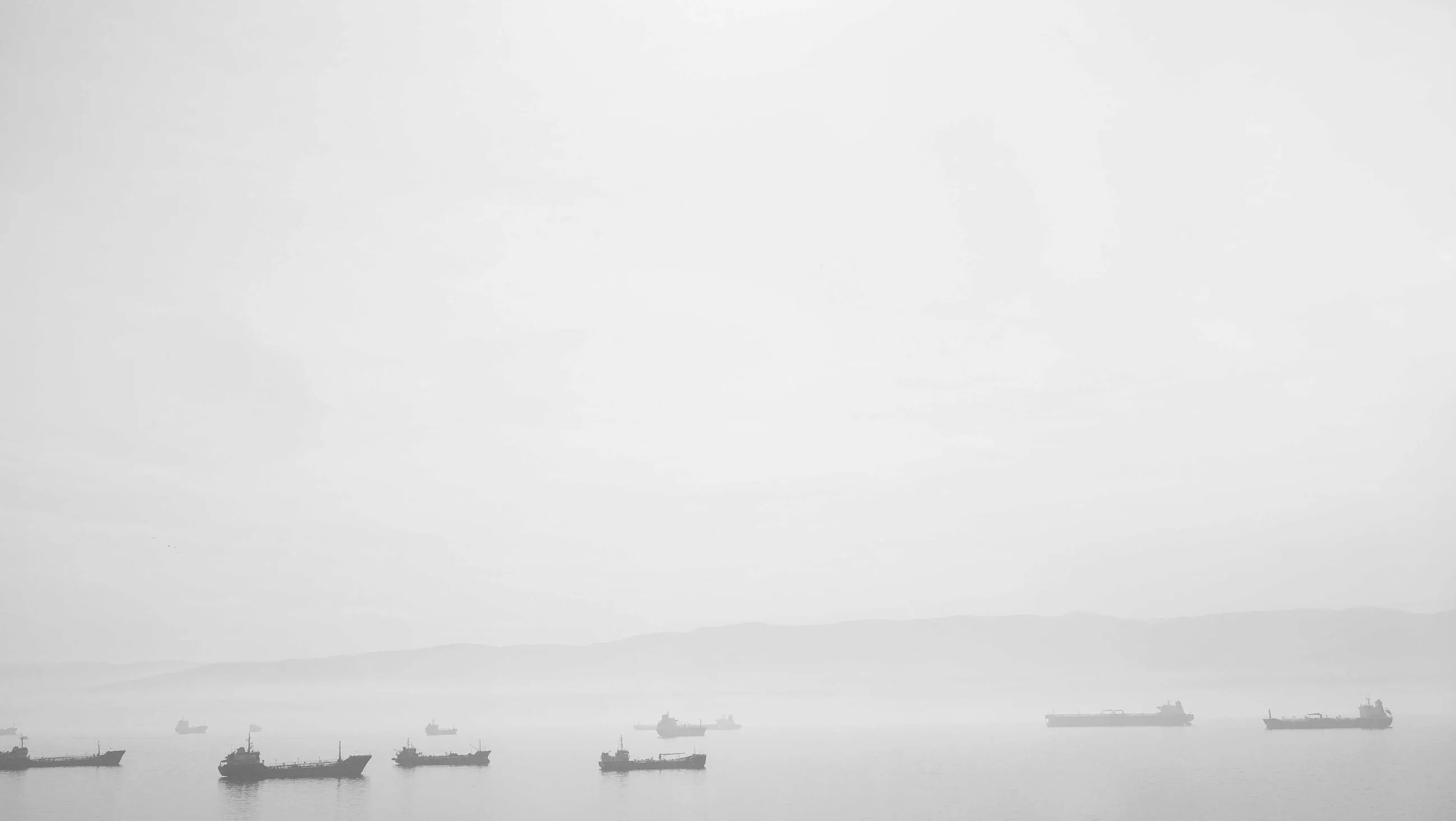 A line of ships at anchor, almost lost in grey fog.