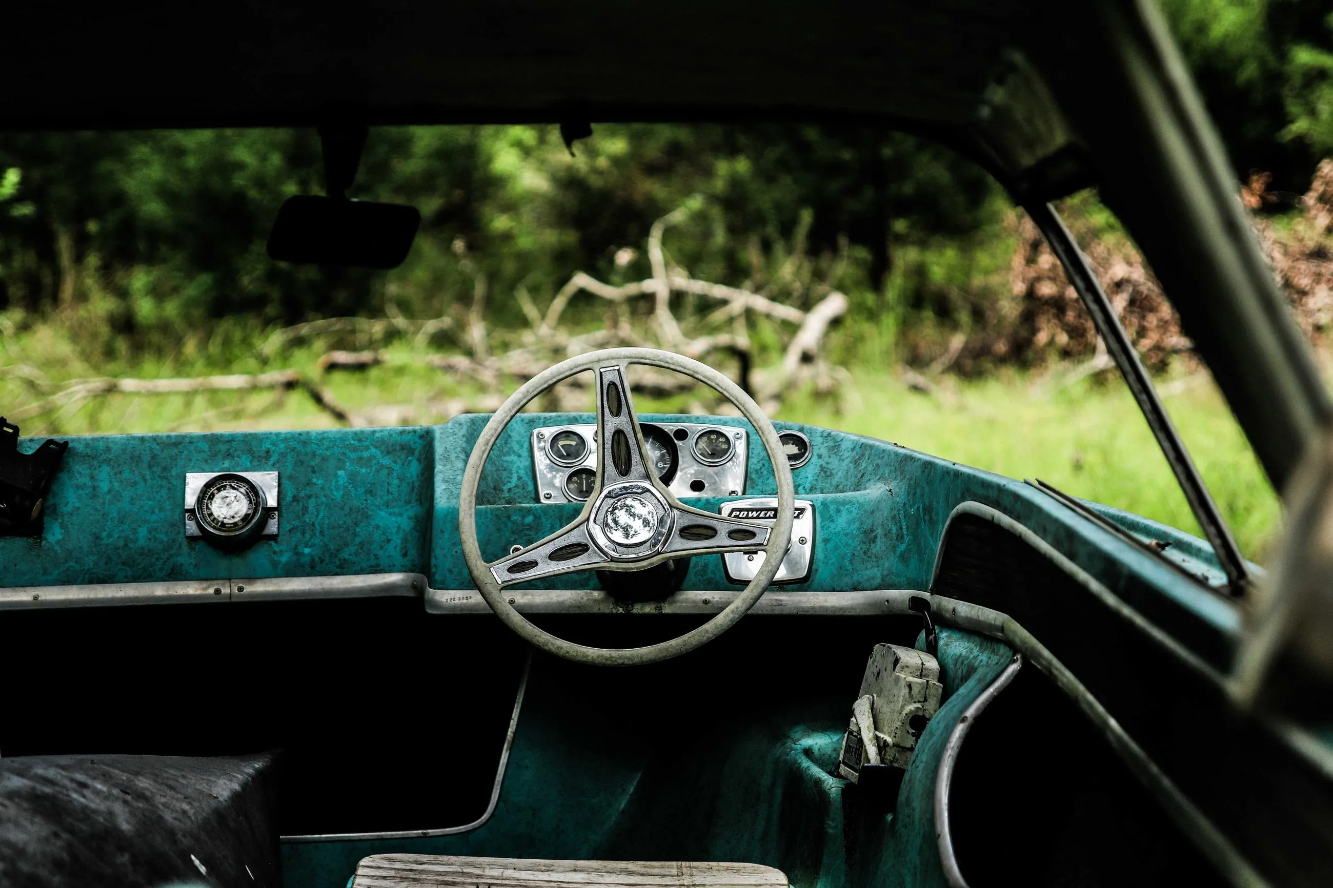 A teal dashboard and a thin steering wheel seen through the open window of an abandoned car.