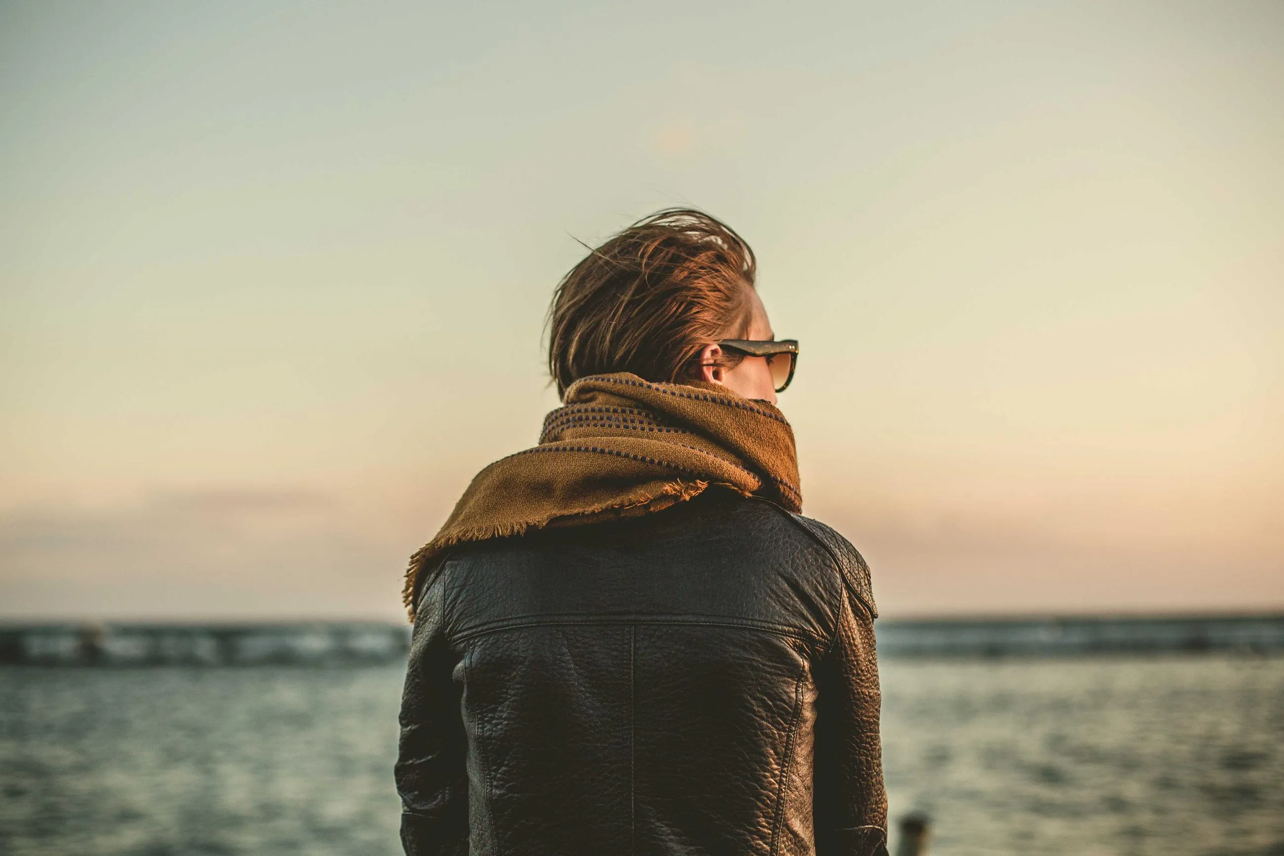 A man in a scarf and leather jacket looks out over the sea at dusk.