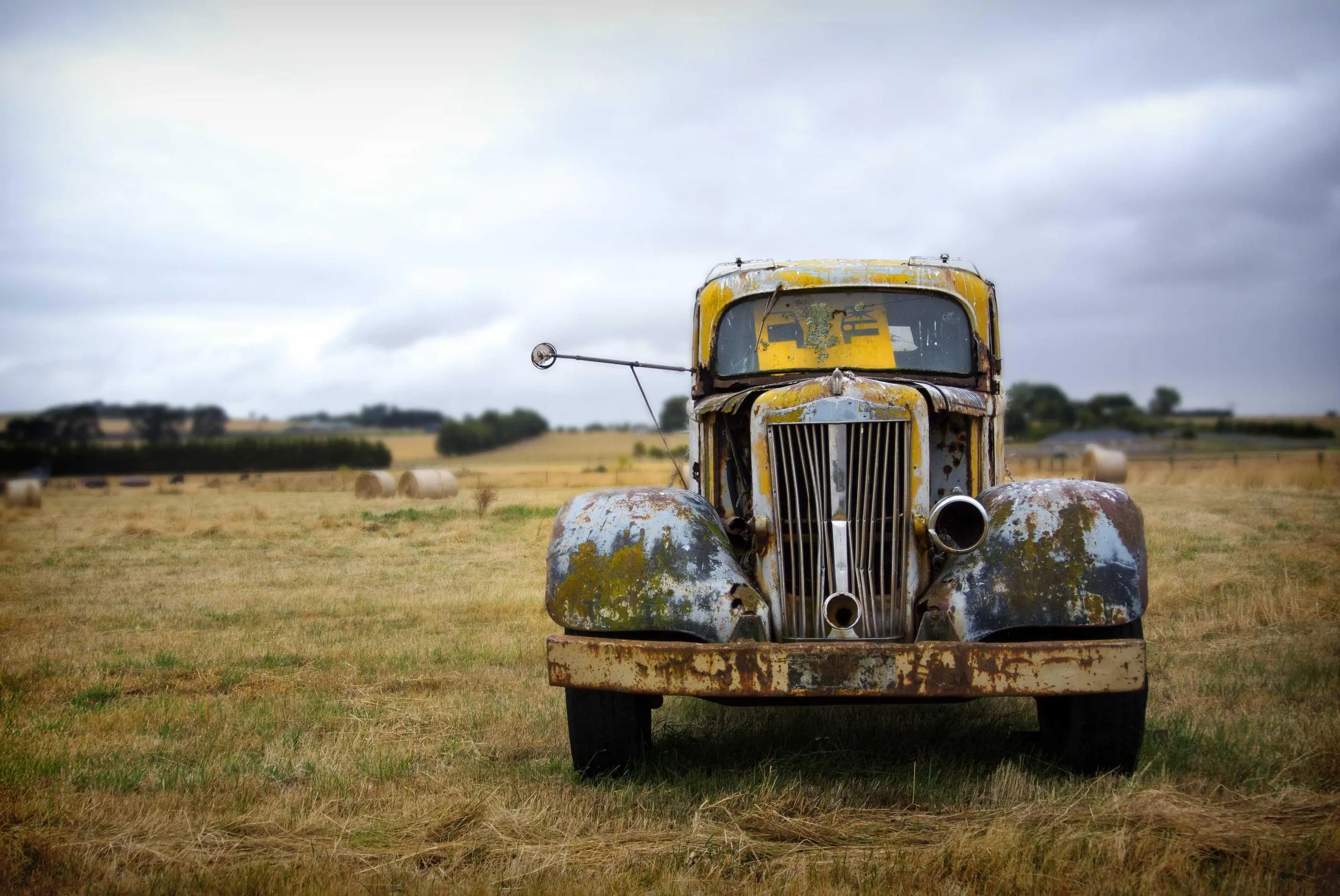 A rusted yellow truck standing alone in a mown field under a heavy sky.