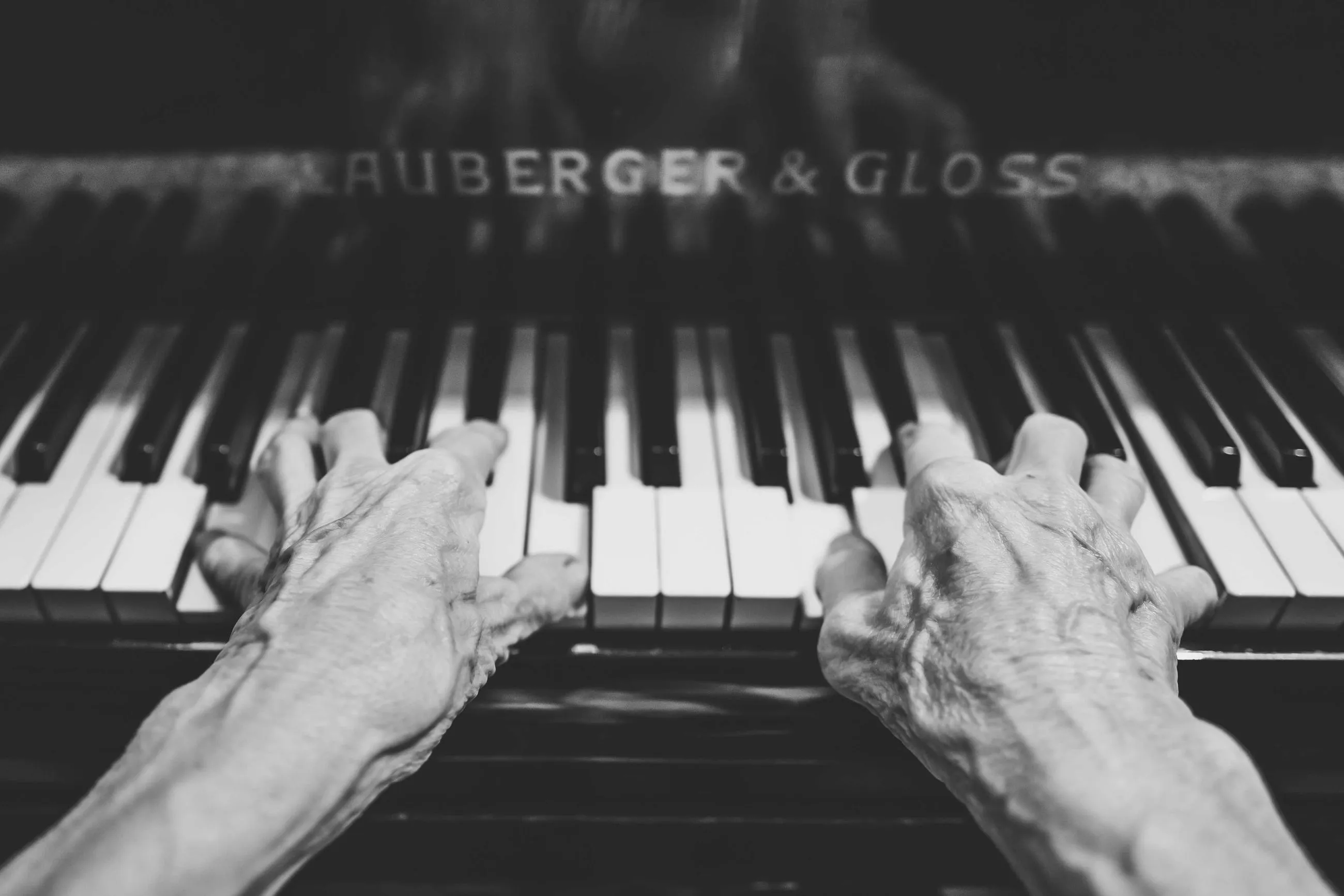 Two old hands resting on the keys of an upright piano, in black and white.