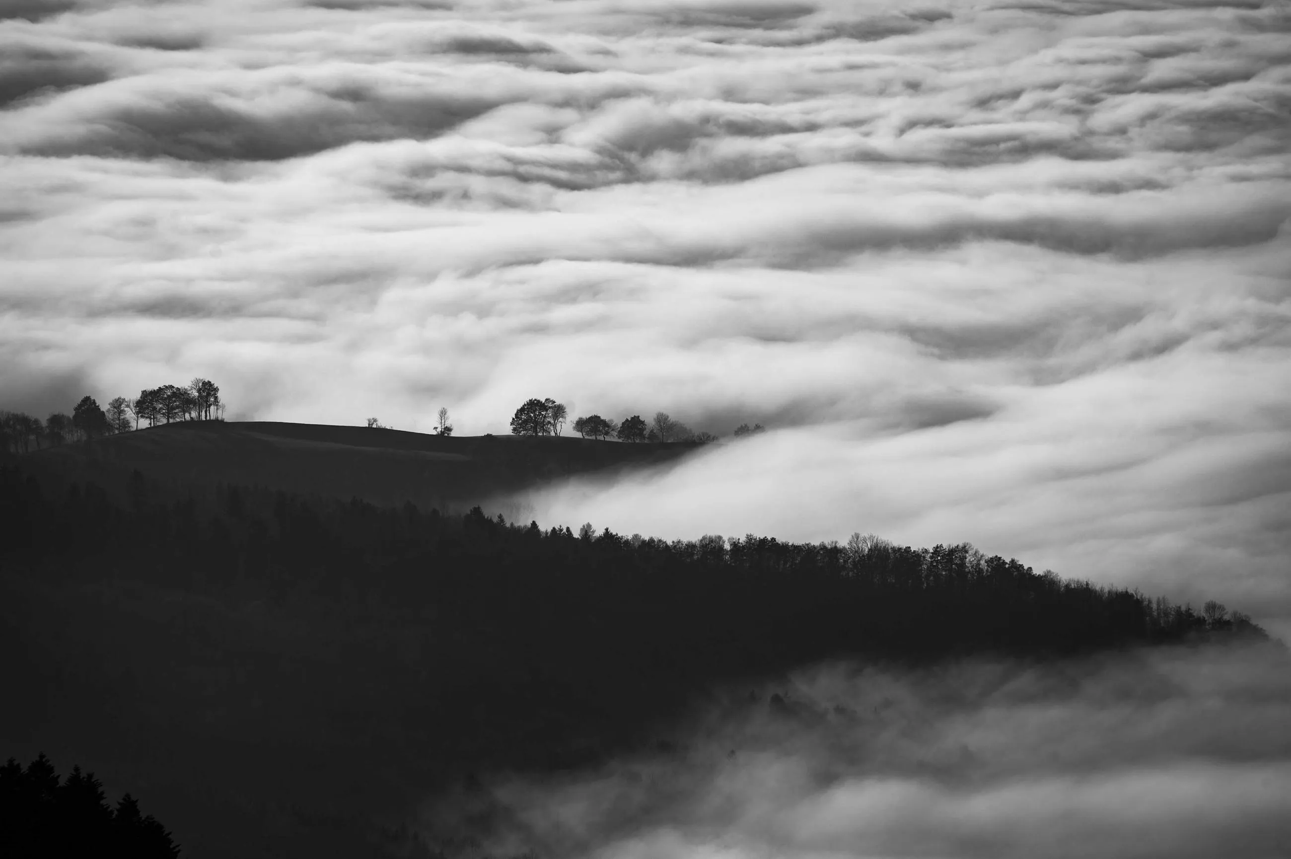 Fog pouring over a dark wooded ridge, in black and white.